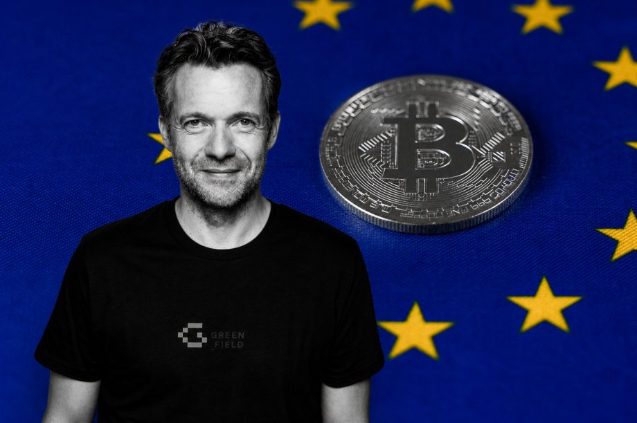 Schwarz-weißes Porträt eines lächelnden Mannes vor einem blauen Hintergrund mit gelben Europasternen; neben ihm liegt eine graue Bitcoin-Münze auf der Flagge der Europa-Union.