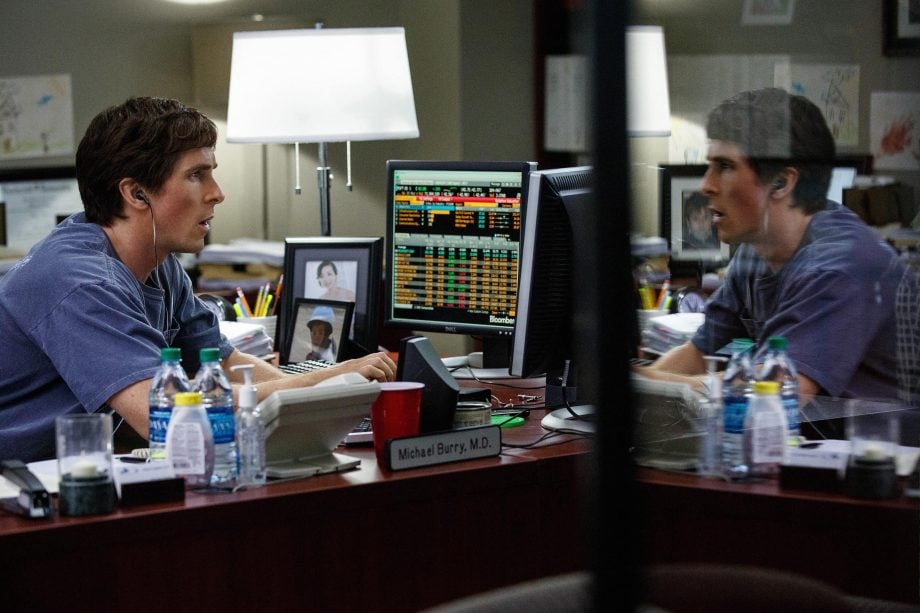 Ein Mann im Kittel, der an Michael Burry erinnert, sitzt an einem Schreibtisch mit Computermonitoren, auf denen Finanzdaten angezeigt werden, und trägt Kopfhörer, in denen sich sein Spiegelbild spiegelt.