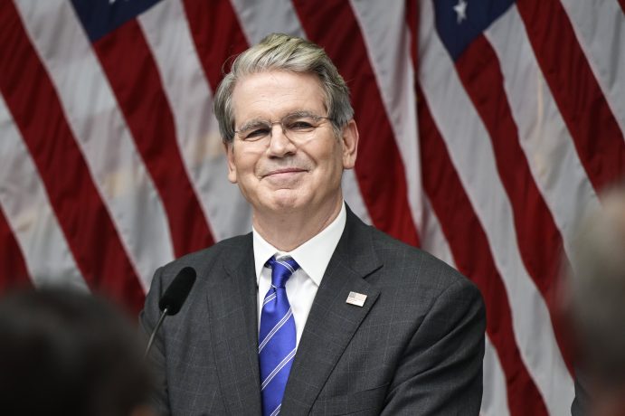 Ein Mann in Anzug und Krawatte (US-Finanzminister Scott Bessent) steht vor mehreren amerikanischen Flaggen und lächelt von einem Podium mit einem Mikrofon, als der US-Finanzminister neue Bitcoin-Regelungen ankündigt.