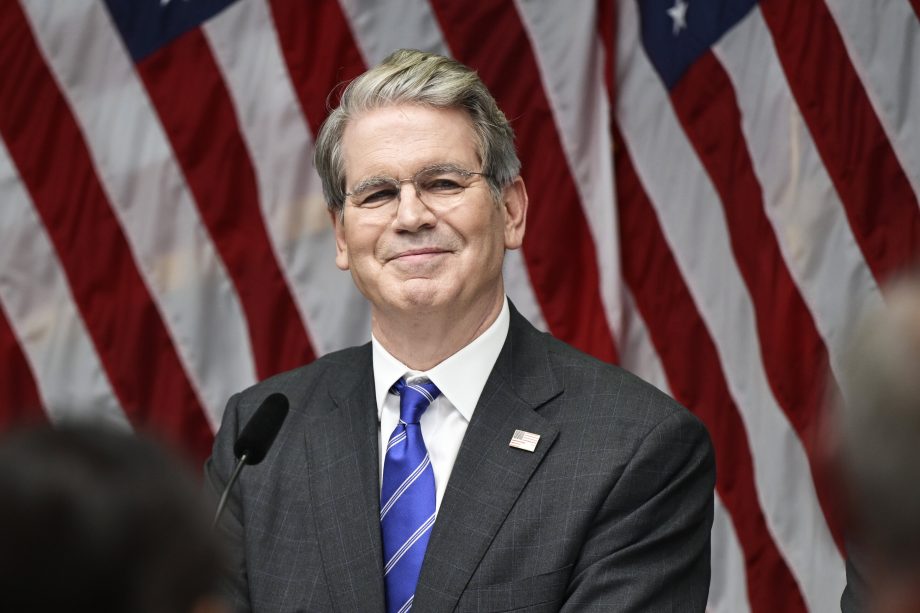 Ein Mann in Anzug und Krawatte (US-Finanzminister Scott Bessent) steht vor mehreren amerikanischen Flaggen und lächelt von einem Podium mit einem Mikrofon, als der US-Finanzminister neue Bitcoin-Regelungen ankündigt.