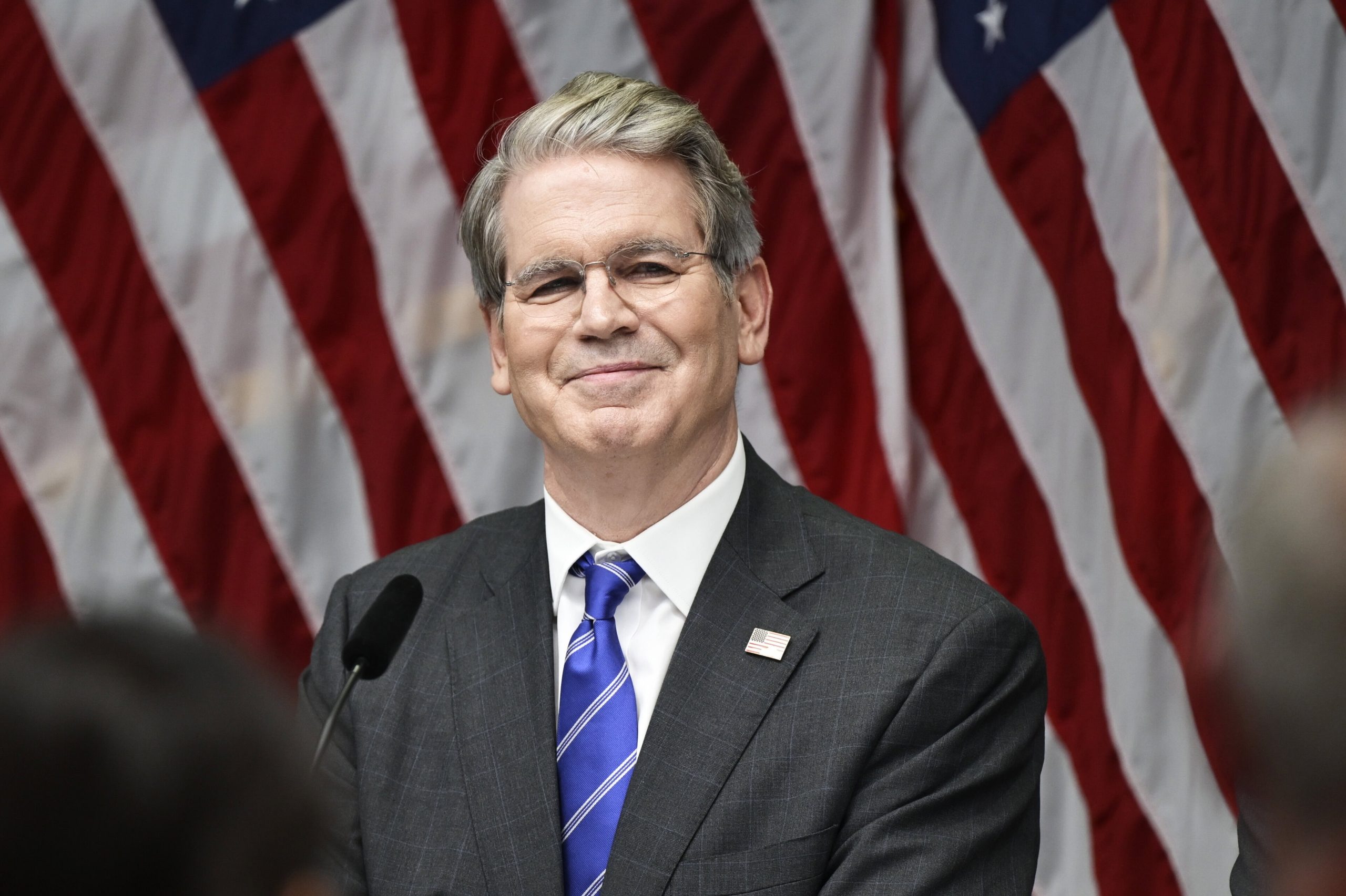 Ein Mann in Anzug und Krawatte (US-Finanzminister Scott Bessent) steht vor mehreren amerikanischen Flaggen und lächelt von einem Podium mit einem Mikrofon, als der US-Finanzminister neue Bitcoin-Regelungen ankündigt.