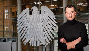 Der Deutsche Bundesadler hängt im Bundestag und rechts sieht man einen Mann mit schwarzem Rollkragenpullover