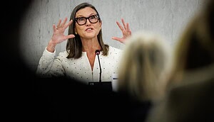 Das Bild zeigt eine Frau, die während einer öffentlichen Veranstaltung oder Pressekonferenz an einem Rednerpult spricht. Sie trägt eine helle, elegant strukturierte Jacke sowie eine markante Brille mit dunklem Rahmen. Ihre schulterlangen Haare sind sorgfältig gestylt, und ihr Auftreten wirkt professionell und selbstbewusst.
