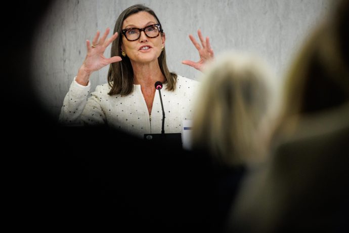 Das Bild zeigt eine Frau, die während einer öffentlichen Veranstaltung oder Pressekonferenz an einem Rednerpult spricht. Sie trägt eine helle, elegant strukturierte Jacke sowie eine markante Brille mit dunklem Rahmen. Ihre schulterlangen Haare sind sorgfältig gestylt, und ihr Auftreten wirkt professionell und selbstbewusst.