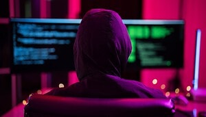 Das Bild zeigt eine Person in einem dunklen Raum, die mit dem Rücken zur Kamera vor mehreren Bildschirmen sitzt. Die Person trägt einen Hoodie, wodurch Gesicht und Identität verborgen bleiben. Die Monitore im Hintergrund zeigen unscharfe Codezeilen oder Datenstrukturen in grün-blauen Farbtönen.