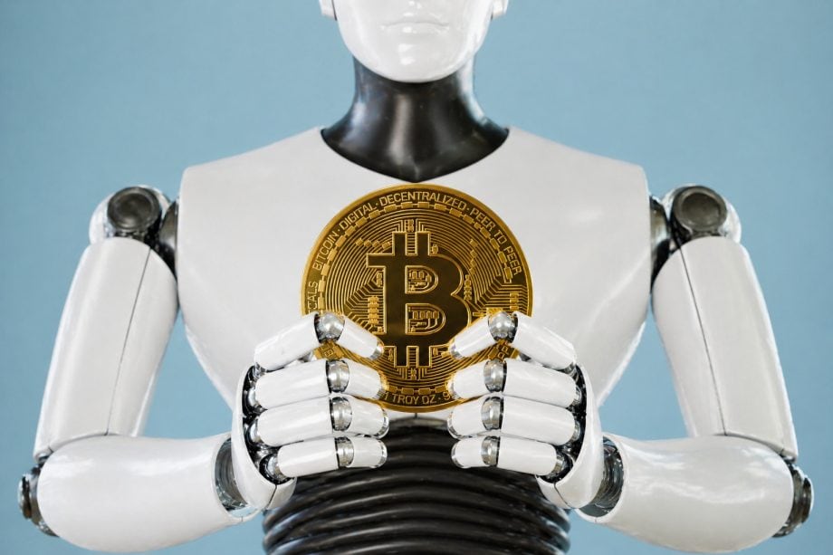 Ein humanoider Roboter hält mit seinen Händen eine Bitcoin-Münze vor eiem hellblauen Hintergrund.