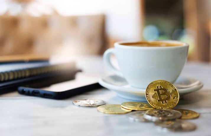 Ein paar Bitcoin-Münzen vor einer Kaffeetasse und einem Smartphone.