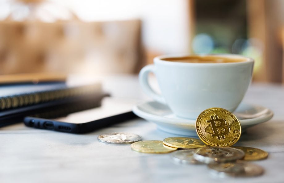 Ein paar Bitcoin-Münzen vor einer Kaffeetasse und einem Smartphone.