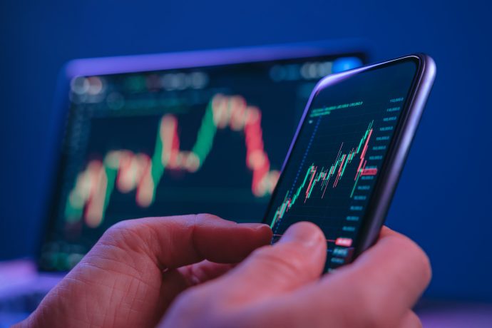 Das Bild zeigt eine Nahaufnahme einer Person, die ein Smartphone in der Hand hält und darauf eine Trading-App betrachtet. Auf dem Display sind farbige Candlestick-Charts zu sehen, die Kursbewegungen an den Finanzmärkten darstellen. Grüne und rote Kerzen symbolisieren steigende und fallende Preise, während Linien und Skalen auf eine detaillierte Marktanalyse hinweisen. Im Hintergrund ist ein weiterer Bildschirm leicht unscharf erkennbar, der ebenfalls ein Kursdiagramm anzeigt. Diese Tiefenunschärfe verstärkt den Fokus auf das Smartphone und unterstreicht die mobile Natur des Tradings. Die dominierenden Blau- und Lilatöne verleihen dem Bild eine moderne, technologische Atmosphäre.