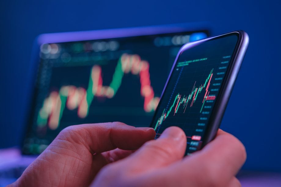 Das Bild zeigt eine Nahaufnahme einer Person, die ein Smartphone in der Hand hält und darauf eine Trading-App betrachtet. Auf dem Display sind farbige Candlestick-Charts zu sehen, die Kursbewegungen an den Finanzmärkten darstellen. Grüne und rote Kerzen symbolisieren steigende und fallende Preise, während Linien und Skalen auf eine detaillierte Marktanalyse hinweisen. Im Hintergrund ist ein weiterer Bildschirm leicht unscharf erkennbar, der ebenfalls ein Kursdiagramm anzeigt. Diese Tiefenunschärfe verstärkt den Fokus auf das Smartphone und unterstreicht die mobile Natur des Tradings. Die dominierenden Blau- und Lilatöne verleihen dem Bild eine moderne, technologische Atmosphäre.