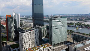 Zu sehen ist die Wiener Skyline mit modernen Büro- und Hochhäusern entlang der Donau. Im Zentrum steht ein markanter, dunkler Glasturm, umgeben von weiteren Gebäuden des Donau-City-Komplexes. Im Hintergrund verlaufen Fluss, Brücken und Teile der Stadtlandschaft unter leicht bewölktem Himmel.