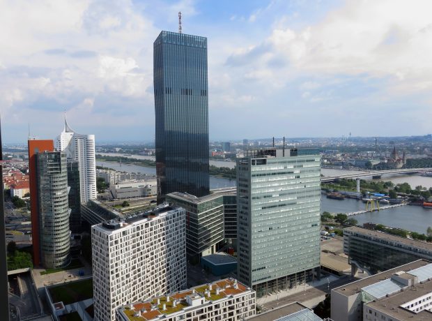 Zu sehen ist die Wiener Skyline mit modernen Büro- und Hochhäusern entlang der Donau. Im Zentrum steht ein markanter, dunkler Glasturm, umgeben von weiteren Gebäuden des Donau-City-Komplexes. Im Hintergrund verlaufen Fluss, Brücken und Teile der Stadtlandschaft unter leicht bewölktem Himmel.