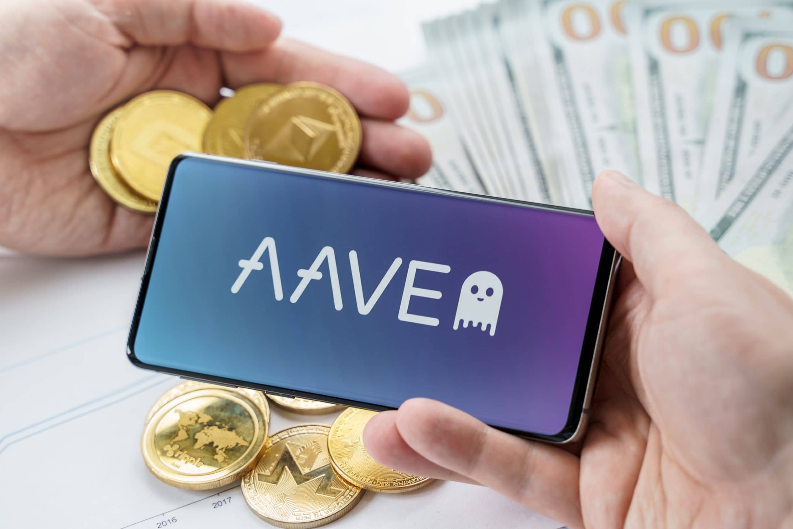 Das Bild zeigt ein Smartphone, auf dessen Display das Logo des DeFi-Protokolls Aave zu sehen ist. Das Gerät wird von zwei Händen gehalten und liegt über mehreren Krypto-Coins, darunter erkennbar Ethereum und andere Token. Im Hintergrund sind unscharf US-Dollar-Banknoten zu sehen. Die Darstellung kombiniert damit visuell die Themen DeFi, Kryptowährungen und Kapital.