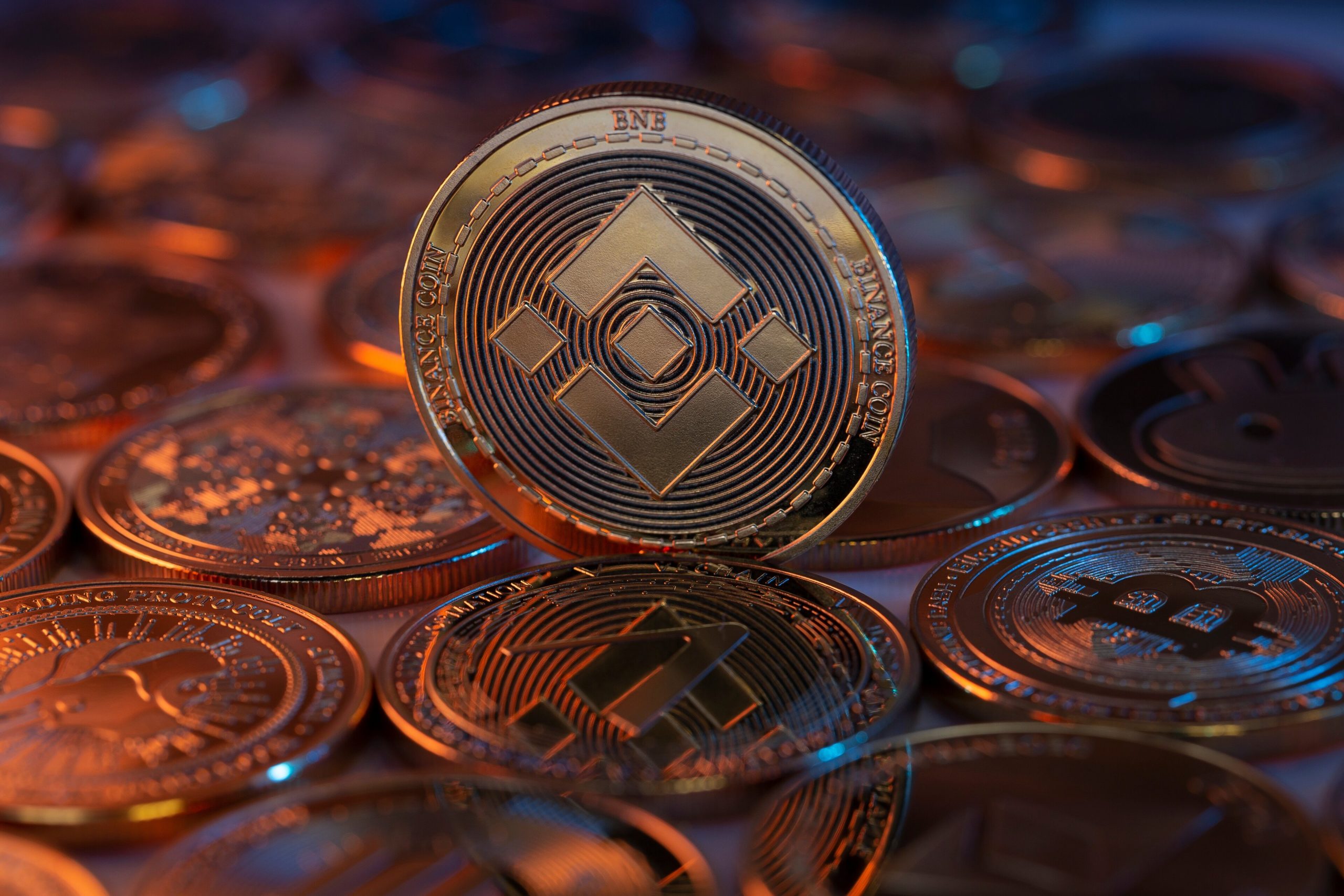 Eine BNB-Münze steht aufrecht zwischen verschiedenen anderen Kryptowährungsmünzen, darunter Bitcoin, und wird von blauem und orangefarbenem Licht beleuchtet.