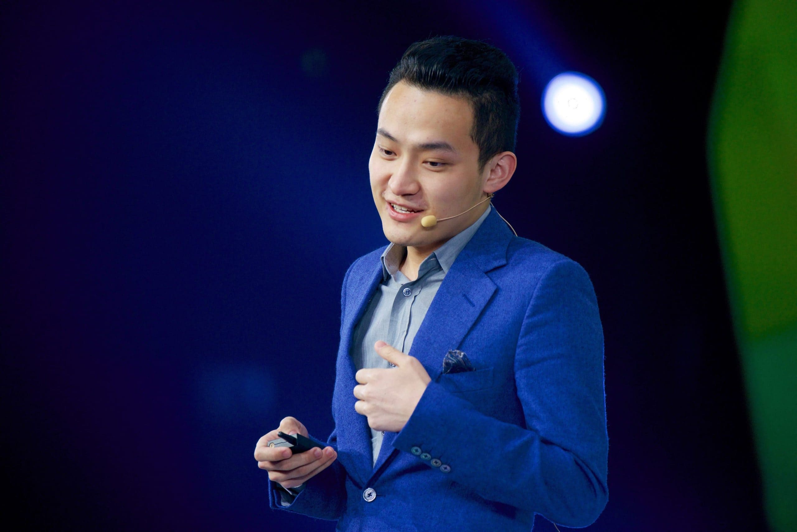 Justin Sun ist elegant in einen blauen Anzug gekleidet, spricht selbstbewusst in ein Headset-Mikrofon und leitet das Publikum auf der schwach beleuchteten Bühne nahtlos mit seinem Klicker an.