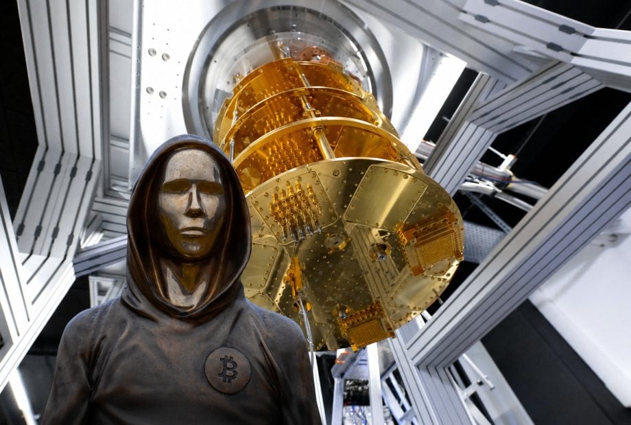 Eine futuristische Szene in einem Hightech-Labor: Im Vordergrund steht eine dunkle, kapuzenartige Bronze-Statue mit maskenhaftem Gesicht und einem Bitcoin-Symbol auf der Brust. Hinter der Figur ragt ein goldfarbener Quantencomputer mit komplexen metallischen Strukturen und Kabeln nach oben. Die Umgebung wirkt steril und modern, mit silbernen Rahmen, weißen Lichtflächen und einer sci-fi-artigen Atmosphäre.