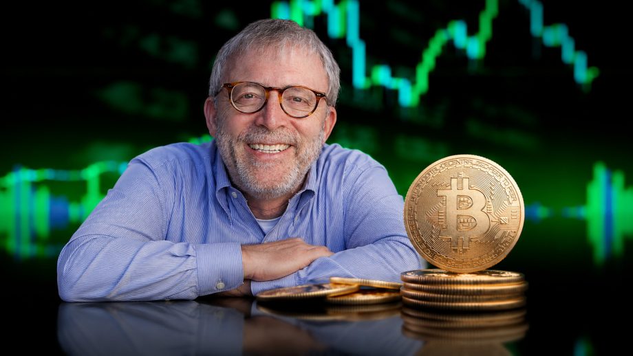 Peter Brandt lächelt in die Kamera und sitzt vor einem Hintergrund mit grünen Bitcoin-Kurscharts. Vor ihm liegen mehrere goldene Bitcoin-Münzen auf einem glänzenden Tisch. Er trägt eine hellblaue Hemdbluse und eine runde Brille.