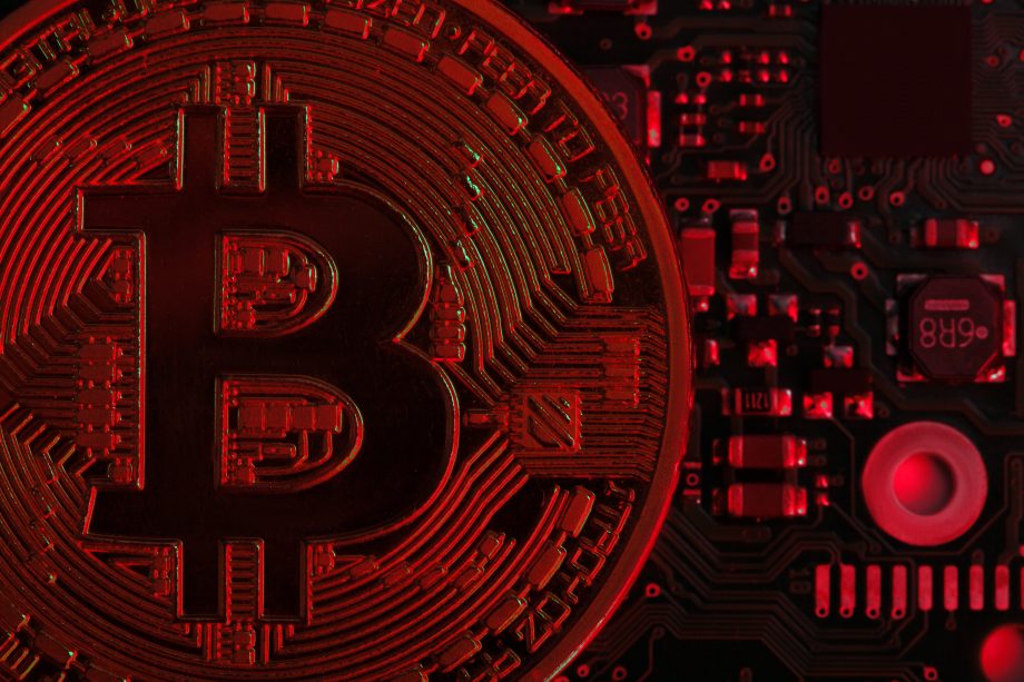 Eine stark vergrößerte Bitcoin-Münze ist vor einer elektronischen Leiterplatte zu sehen, wobei die Szene in rotes Licht getaucht ist und eine negative oder angespannte Marktsituation symbolisiert.