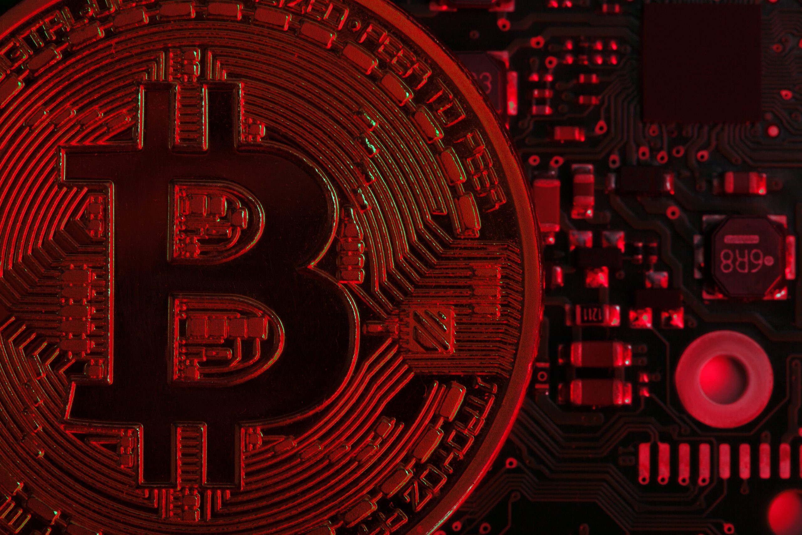 Eine stark vergrößerte Bitcoin-Münze ist vor einer elektronischen Leiterplatte zu sehen, wobei die Szene in rotes Licht getaucht ist und eine negative oder angespannte Marktsituation symbolisiert.