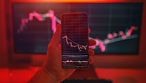 Eine Hand hält ein Smartphone, auf dessen Display ein fallender Kursverlauf mit Candlestick-Chart zu sehen ist. Im Hintergrund zeigt ein großer Monitor ebenfalls einen rückläufigen Markt, während die Szene in rotes Licht getaucht ist und eine angespannte Marktsituation visualisiert.