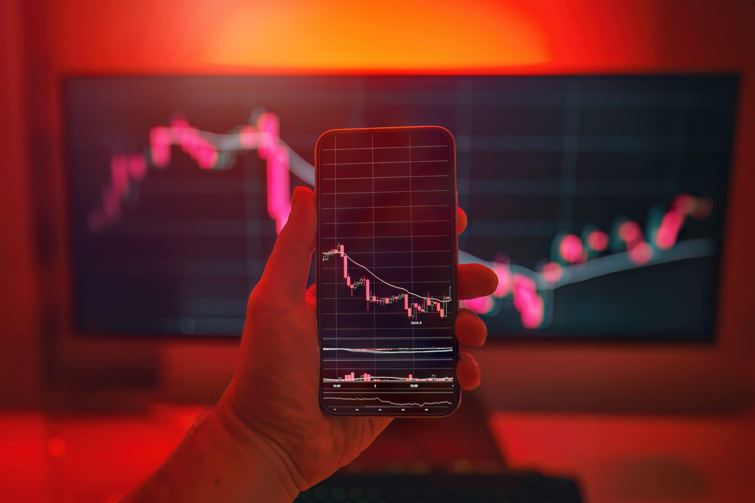 Eine Hand hält ein Smartphone, auf dessen Display ein fallender Kursverlauf mit Candlestick-Chart zu sehen ist. Im Hintergrund zeigt ein großer Monitor ebenfalls einen rückläufigen Markt, während die Szene in rotes Licht getaucht ist und eine angespannte Marktsituation visualisiert.