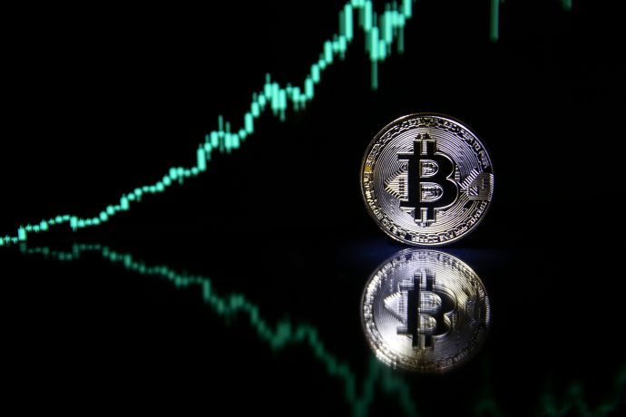 Eine Bitcoin-Münze steht im Vordergrund vor einem dunklen Hintergrund, während im Hintergrund ein aufwärts gerichteter Kurschart zu sehen ist, der sich in der Oberfläche spiegelt.
