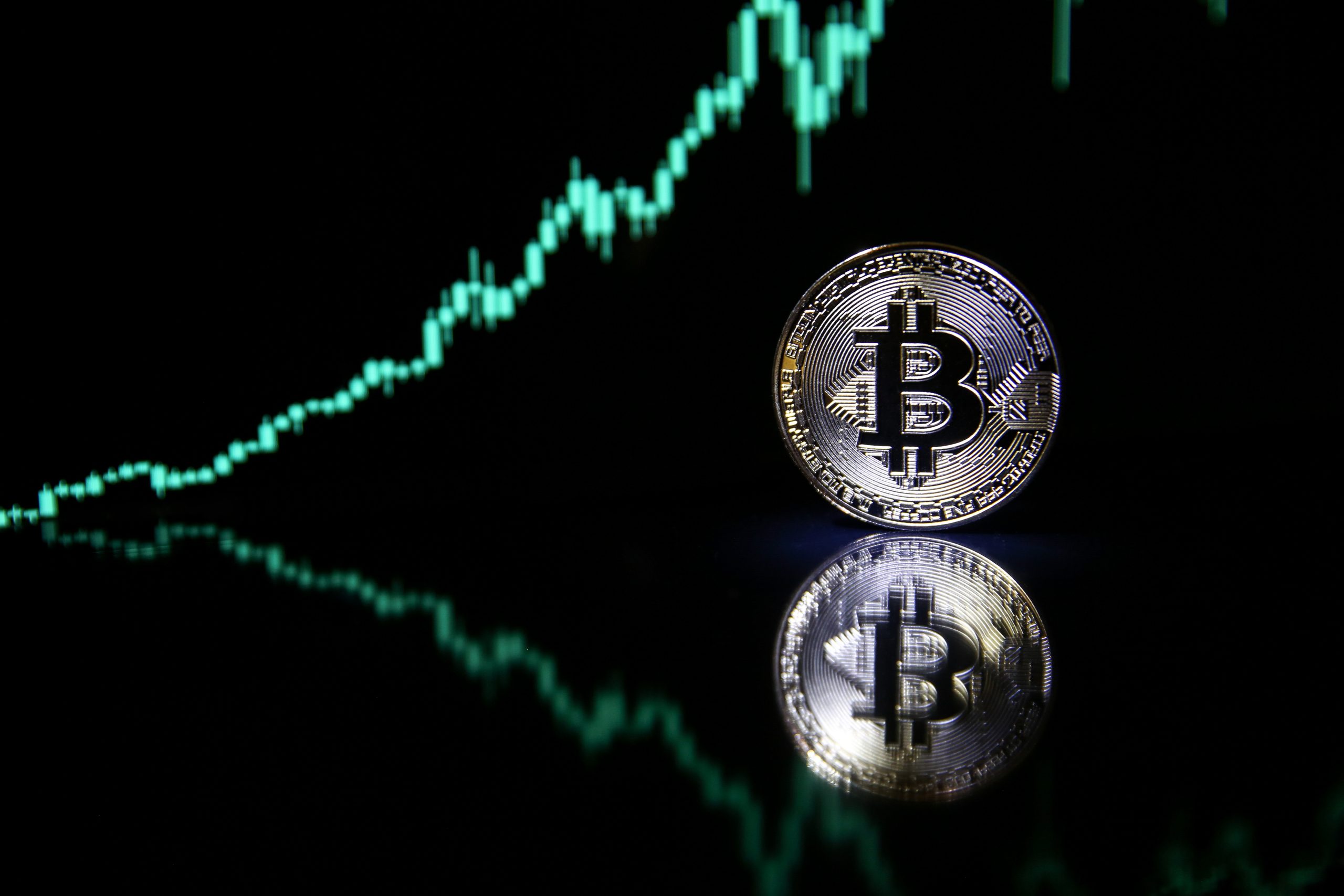 Eine Bitcoin-Münze steht im Vordergrund vor einem dunklen Hintergrund, während im Hintergrund ein aufwärts gerichteter Kurschart zu sehen ist, der sich in der Oberfläche spiegelt.
