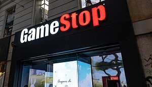 Das Bild zeigt eine GameStop-Filiale in einer Stadtumgebung. Im Mittelpunkt steht das beleuchtete Logo des Unternehmens über dem Eingang, wobei „Game“ in Weiß und „Stop“ in Rot dargestellt ist. Unter dem Schriftzug befindet sich ein Schaufenster mit digitalen Displays, auf denen bunte, spielbezogene Motive zu sehen sind. Im Hintergrund ist die Fassade eines Gebäudes mit Fenstern erkennbar.