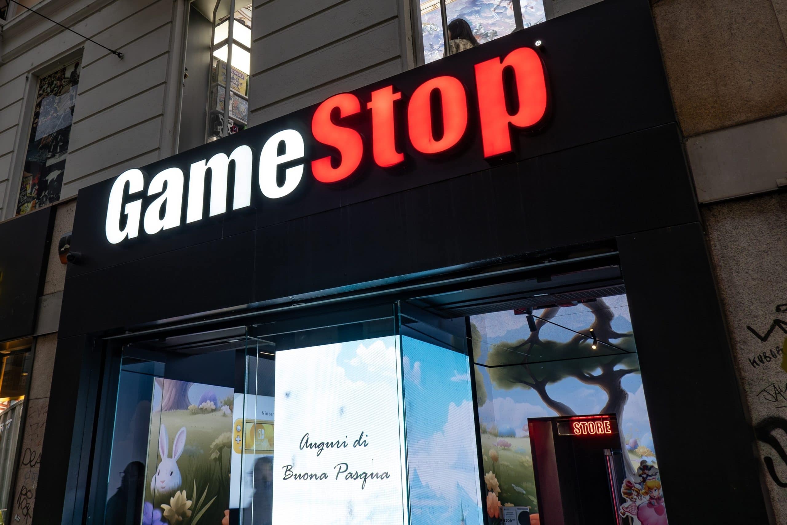 Das Bild zeigt eine GameStop-Filiale in einer Stadtumgebung. Im Mittelpunkt steht das beleuchtete Logo des Unternehmens über dem Eingang, wobei „Game“ in Weiß und „Stop“ in Rot dargestellt ist. Unter dem Schriftzug befindet sich ein Schaufenster mit digitalen Displays, auf denen bunte, spielbezogene Motive zu sehen sind. Im Hintergrund ist die Fassade eines Gebäudes mit Fenstern erkennbar.