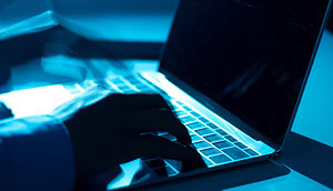 Zu sehen ist eine Nahaufnahme von Händen, die auf einer Laptop-Tastatur tippen. Die Szene ist in kühles, bläuliches Licht getaucht, wodurch eine technisch-düstere Atmosphäre entsteht, die an Cybersecurity oder digitale Angriffe erinnert.