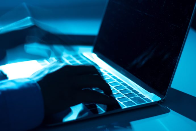 Zu sehen ist eine Nahaufnahme von Händen, die auf einer Laptop-Tastatur tippen. Die Szene ist in kühles, bläuliches Licht getaucht, wodurch eine technisch-düstere Atmosphäre entsteht, die an Cybersecurity oder digitale Angriffe erinnert.