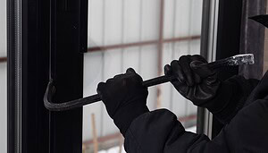 Eine vermummte Person mit schwarzen Handschuhen versucht mit einem Brecheisen eine Tür oder ein Fenster aufzuhebeln. Das Bild symbolisiert physische Einbrüche und Gewaltkriminalität im Zusammenhang mit Krypto-Überfällen.
