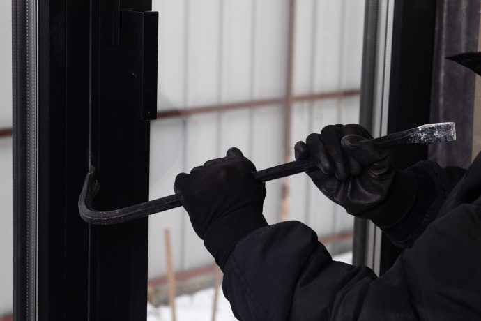 Eine vermummte Person mit schwarzen Handschuhen versucht mit einem Brecheisen eine Tür oder ein Fenster aufzuhebeln. Das Bild symbolisiert physische Einbrüche und Gewaltkriminalität im Zusammenhang mit Krypto-Überfällen.