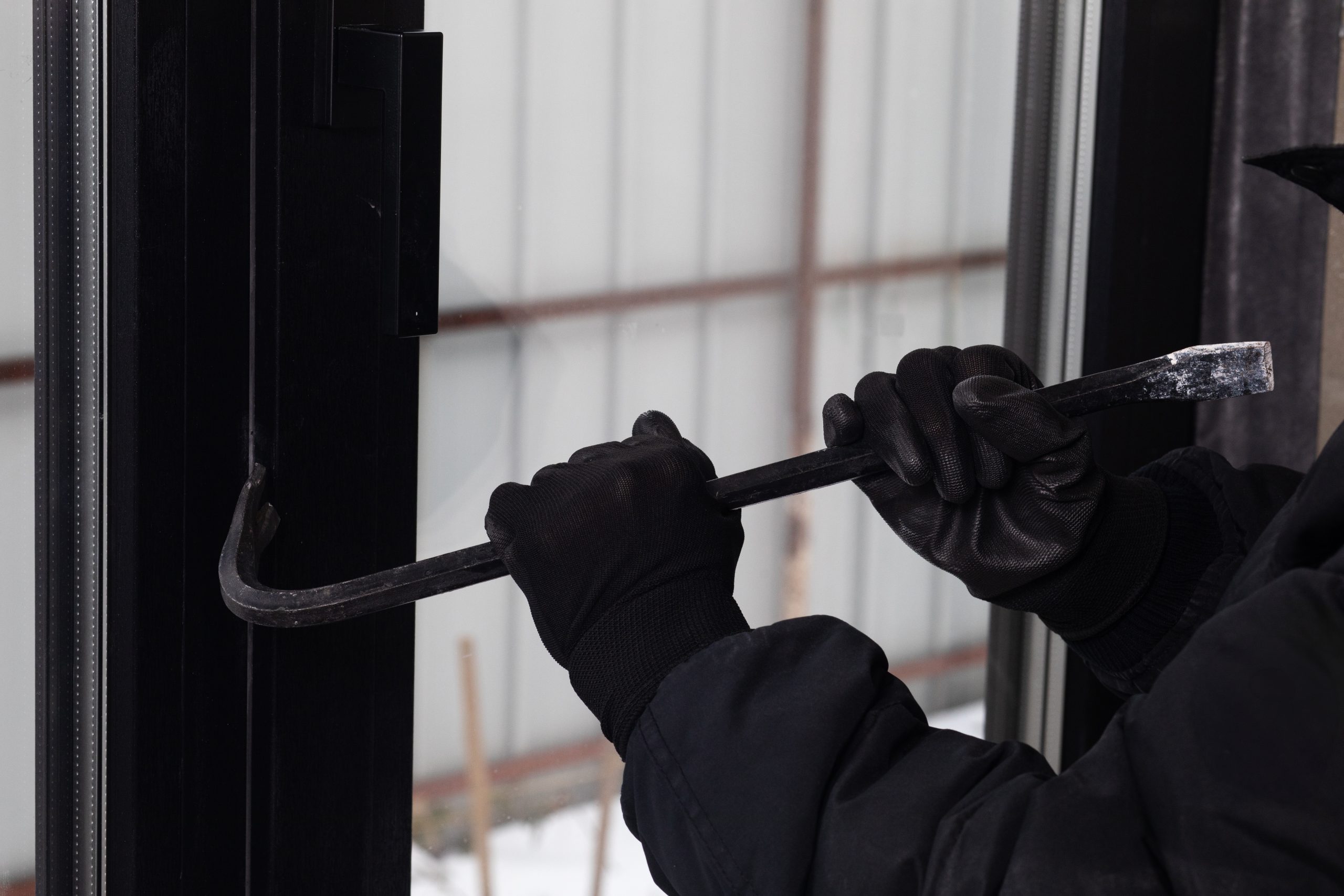 Eine vermummte Person mit schwarzen Handschuhen versucht mit einem Brecheisen eine Tür oder ein Fenster aufzuhebeln. Das Bild symbolisiert physische Einbrüche und Gewaltkriminalität im Zusammenhang mit Krypto-Überfällen.