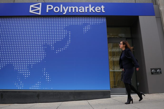 Zu sehen ist die Außenfassade eines Gebäudes mit einem großen „Polymarket“-Schriftzug. Eine Frau geht an dem Eingang vorbei, während im Hintergrund eine stilisierte Weltkarte auf einer blauen Wand dargestellt ist.
