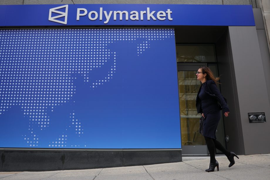 Zu sehen ist die Außenfassade eines Gebäudes mit einem großen „Polymarket“-Schriftzug. Eine Frau geht an dem Eingang vorbei, während im Hintergrund eine stilisierte Weltkarte auf einer blauen Wand dargestellt ist.