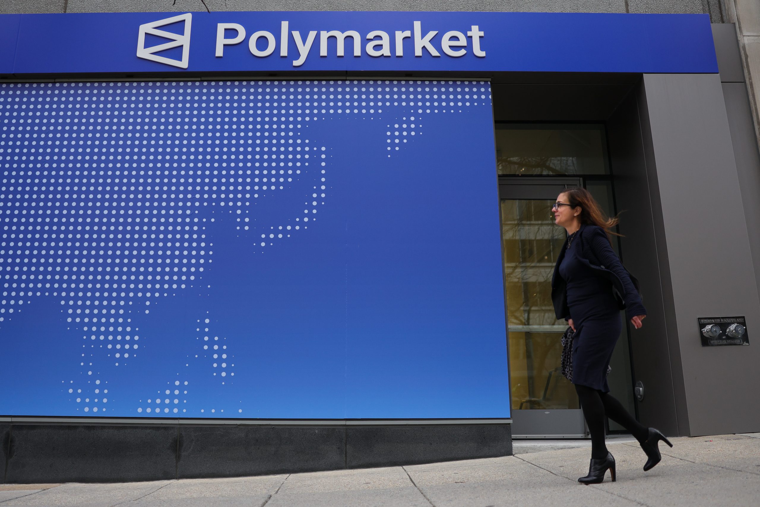 Zu sehen ist die Außenfassade eines Gebäudes mit einem großen „Polymarket“-Schriftzug. Eine Frau geht an dem Eingang vorbei, während im Hintergrund eine stilisierte Weltkarte auf einer blauen Wand dargestellt ist.
