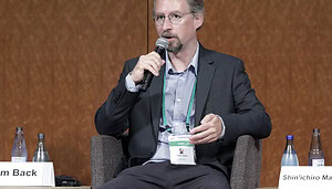 Das Bild zeigt Adam Back auf einer Bühne mit einem Anzug und Mikrofon, wie er vielleicht über Bitcoin und Satoshi Nakamoto spricht.