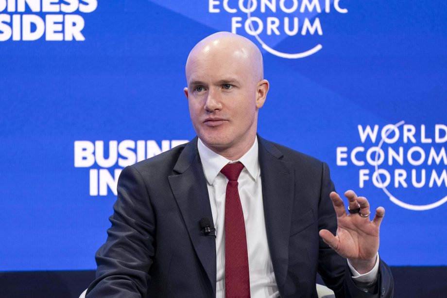 Ein Mann im Anzug und mit roter Krawatte spricht während einer Podiumsdiskussion auf dem Weltwirtschaftsforum, im Hintergrund sind blaue Banner und das Coinbase-Logo zu sehen.