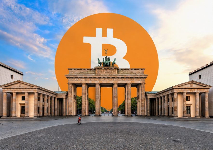 Das Brandenburger Tor in Berlin bei Sonnenaufgang mit leerem Pariser Platz. Hinter dem historischen Wahrzeichen erhebt sich ein überdimensionales orangefarbenes Bitcoin-Logo, das den Himmel dominiert und das Tor optisch in seinen Schatten stellt. Die Kombination aus klassischer Architektur und Kryptowährung symbolisiert die wachsende Bedeutung von Bitcoin in Berlin und Deutschland.