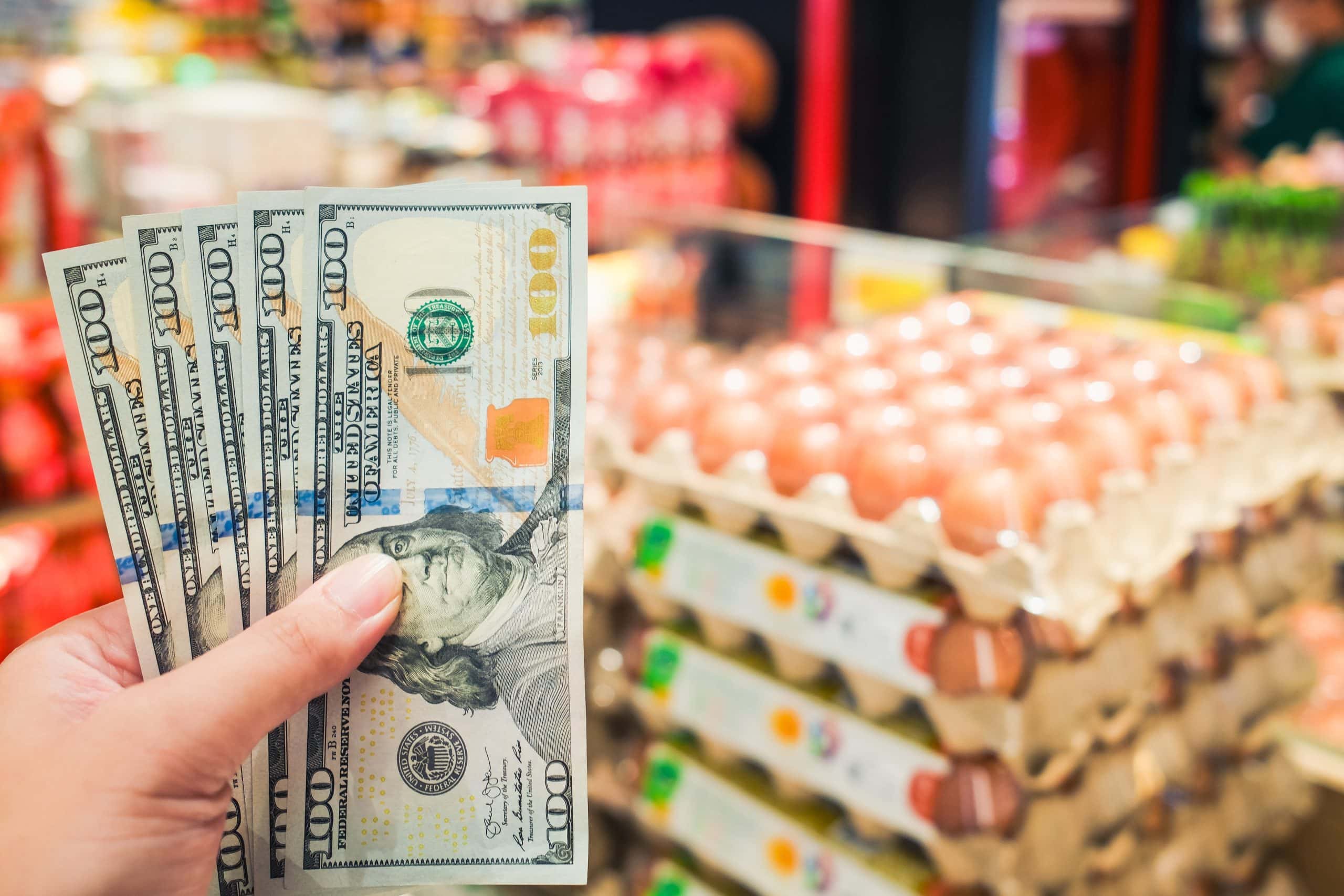 Das Bild zeigt die Hand mit Dollarscheinen und im Hintergrund sind Eier zu sehen.