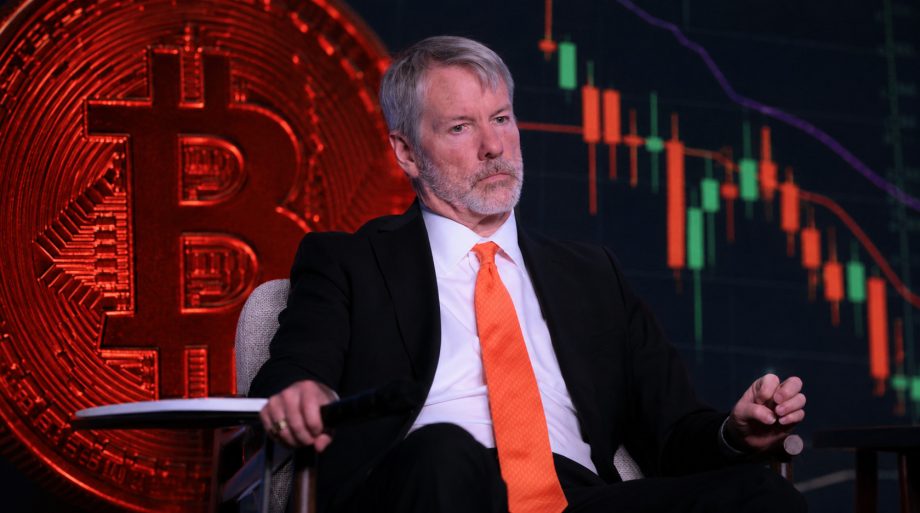 Michael Saylor sitzt in einem Sessel vor einem Hintergrund aus einer großen roten Bitcoin-Münze und einem Kurschart mit roten und grünen Candlesticks. Er trägt einen dunklen Anzug, ein weißes Hemd und eine orangefarbene Krawatte. Sein ernster Blick und die Darstellung des volatilen Kryptomarkts im Hintergrund vermitteln eine angespannte, finanzmarktbezogene Atmosphäre. Die Bildmontage verbindet die Person visuell mit dem Thema Bitcoin und Kryptowährungen.