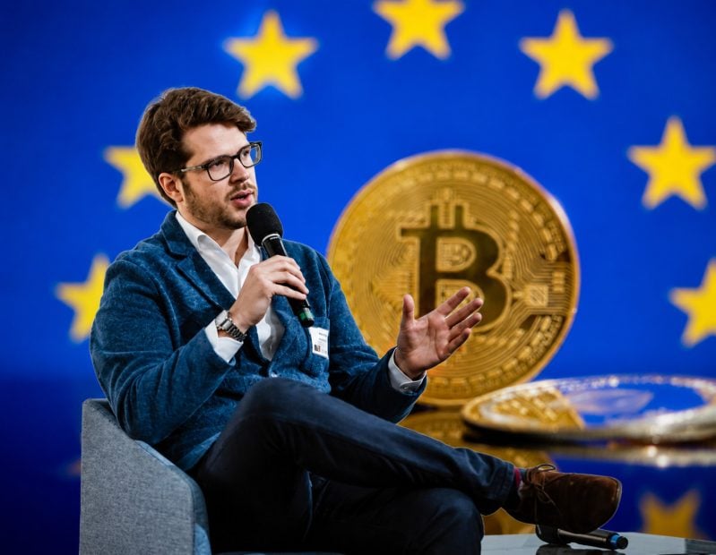 Robert Kopitsch spricht auf einer Konferenz über Bitcoin und die europäische Kryptoregulierung vor einem Hintergrund mit EU-Sternen und Bitcoin-Symbol.