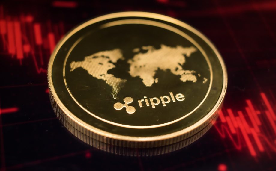 Eine goldene XRP-Münze mit dem Ripple-Logo und einer Weltkarte ruht auf einer dunklen Oberfläche, die von roten Finanzdiagrammlinien beleuchtet wird.