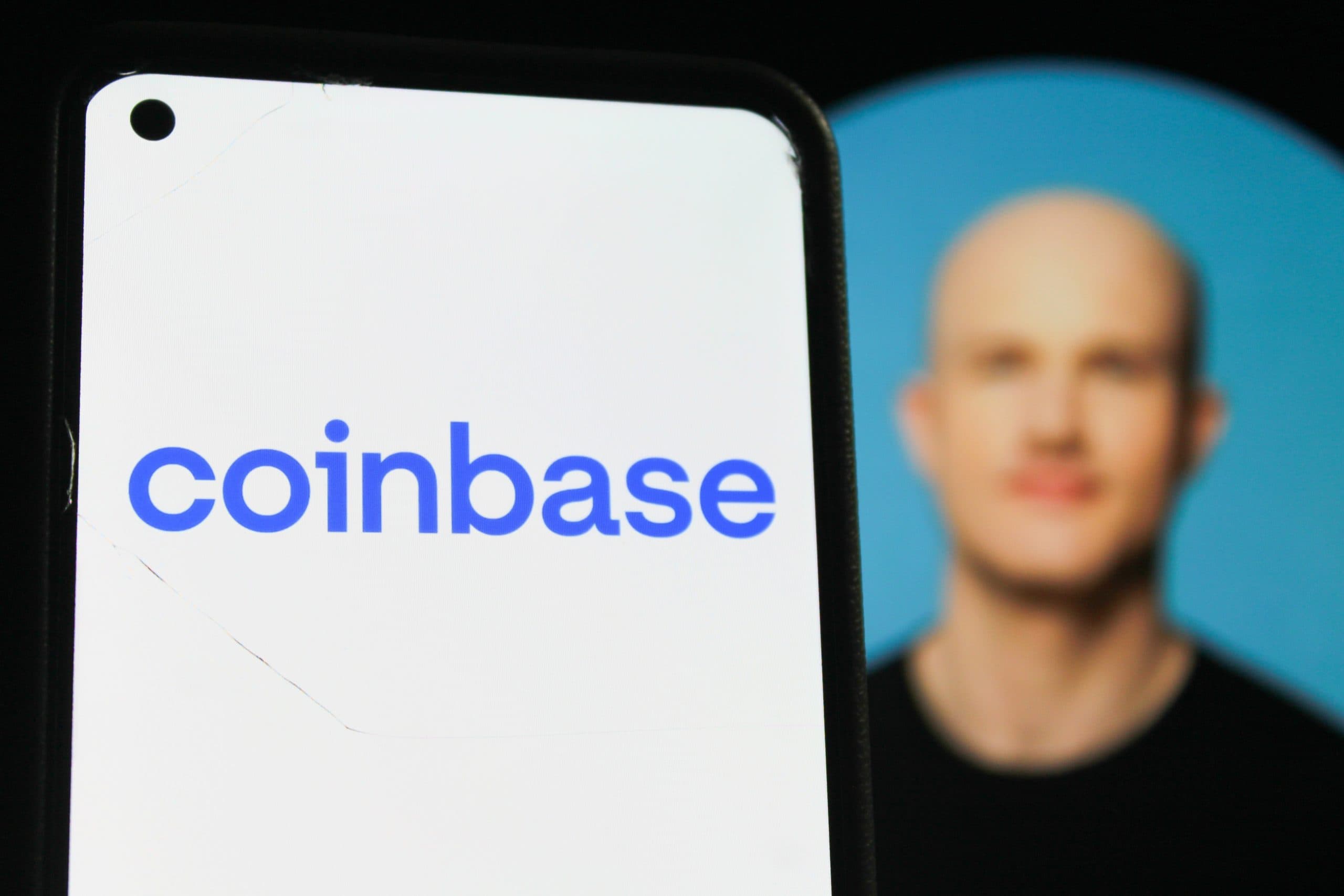 Das Bild zeigt im Vordergrund ein Smartphone mit dem Coinbase-Logo auf dem Display. Im unscharfen Hintergrund ist Coinbase-CEO Brian Armstrong vor einem blauen Hintergrund zu sehen. Durch die geringe Tiefenschärfe liegt der Fokus klar auf dem Unternehmenslogo.