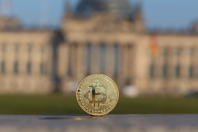 Eine Bitcoin-Münze vor einem verschwommenen Bild des Deutschen Bundestags.