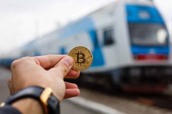 Das Bild zeigt eine Bitcoin-Münze vor einem Zug