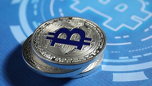 Das Bild zeigt zwei übereinanderliegende physische Bitcoin-Münzen in Nahaufnahme auf blauem Hintergrund. Im Zentrum der oberen Münze befindet sich das stilisierte Bitcoin-Symbol in dunklem Blau. Im Hintergrund ist unscharf eine grafische Darstellung des Bitcoin-Logos zu erkennen, die ebenfalls in Blautönen gehalten ist.