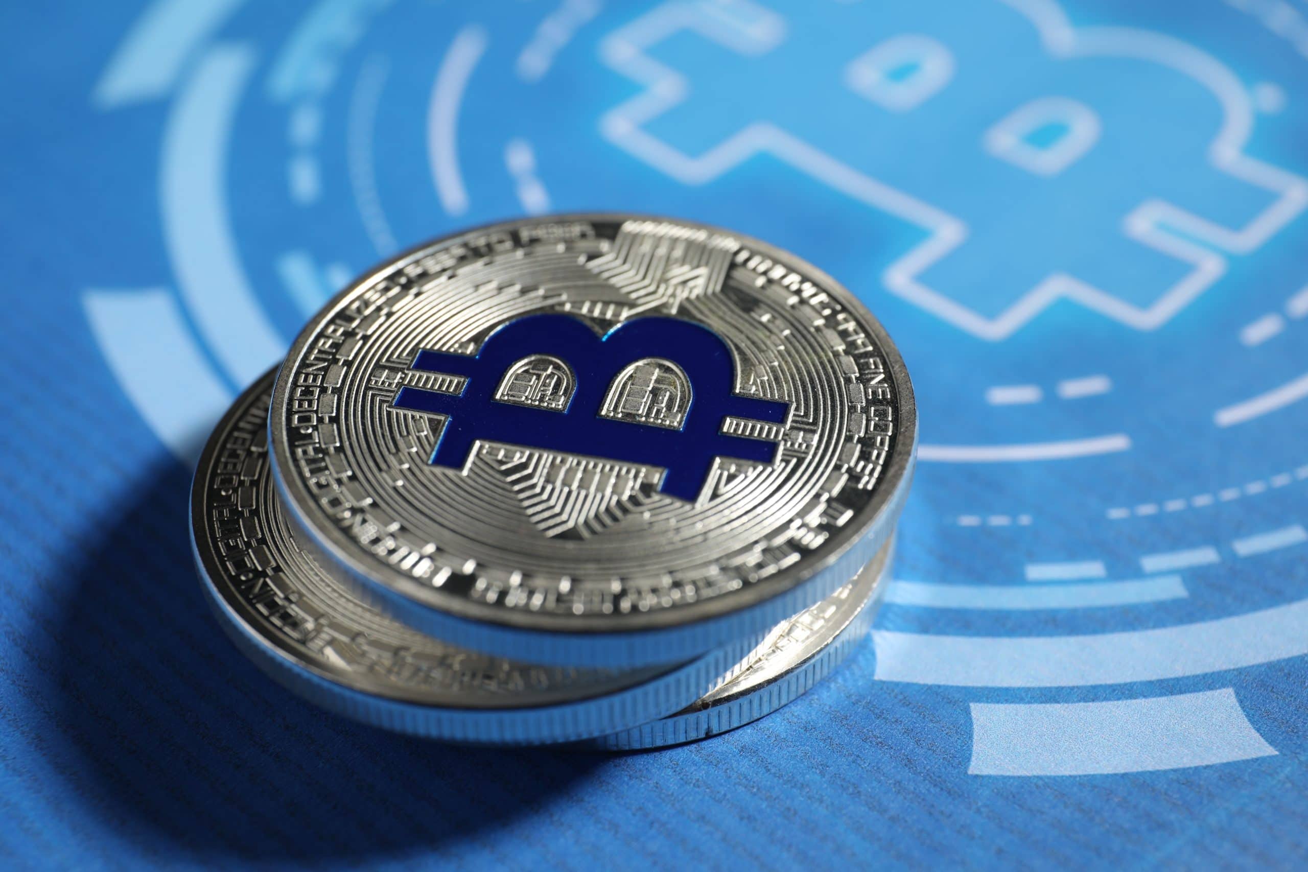 Das Bild zeigt zwei übereinanderliegende physische Bitcoin-Münzen in Nahaufnahme auf blauem Hintergrund. Im Zentrum der oberen Münze befindet sich das stilisierte Bitcoin-Symbol in dunklem Blau. Im Hintergrund ist unscharf eine grafische Darstellung des Bitcoin-Logos zu erkennen, die ebenfalls in Blautönen gehalten ist.