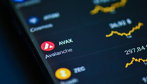 Das Bild zeigt die Kryptowährung Avalanche (AVAX) auf dem Display eines Smartphones oder Tablets. Im Fokus stehen das rote Avalanche-Logo und die Bezeichnung "AVAX". Im Hintergrund sind unscharfe Kurscharts und weitere Kryptowährungen zu erkennen, darunter Bitcoin. Die Aufnahme vermittelt den Eindruck einer Handels- oder Portfolio-App.